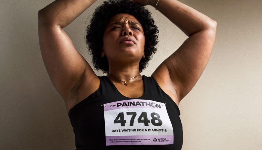 Mujer con expresión de dolor levantando los brazos con los ojos cerrados, y un dorsal de la campaña Painathon en la camiseta.
