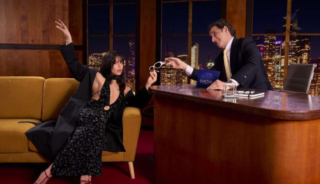 Nathy Peluso en el late show ficticio creado por Melle, cogiendo unas gafas de sol que le tiende el presentador del programa.