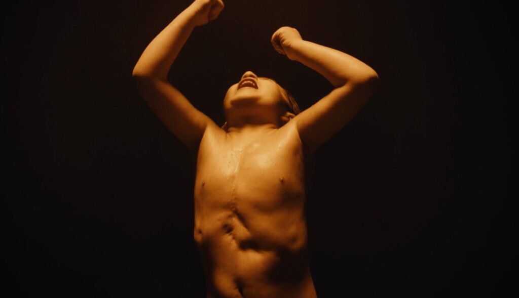 Niño sin camiseta gritando con los brazos en alto sobre un fondo negro