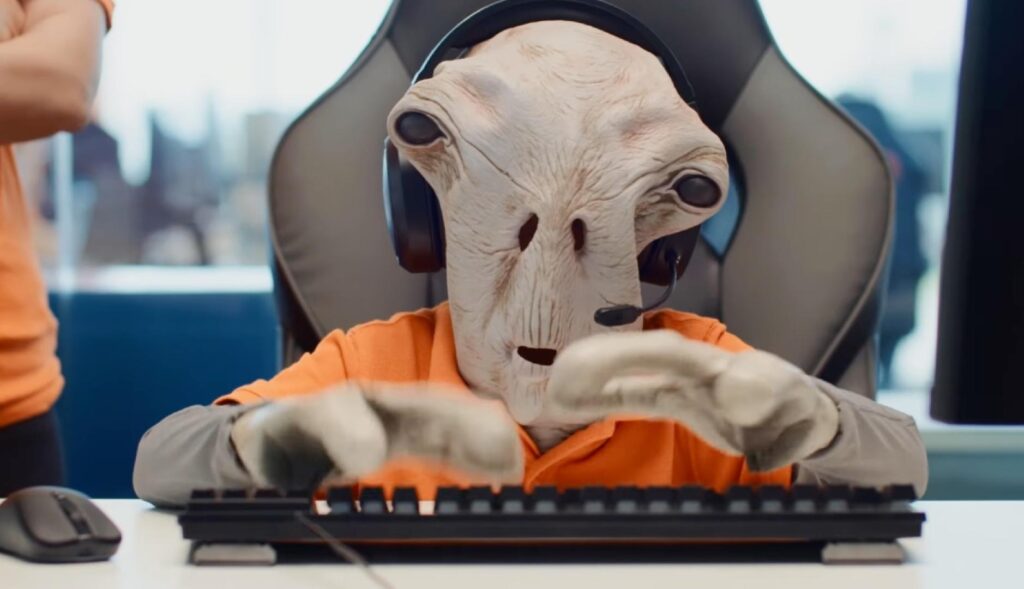 un marciano vestido con el uniforme de pccomponentes sentado delante del ordenador con unos auriculares de centralita