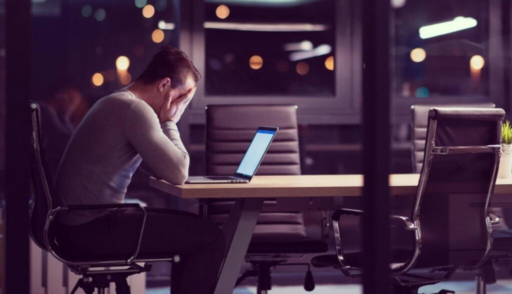 un hombre de noche en una sala de reuniones, él solo se echa las manos a la cara en un gesto de desesperación frente al portátil