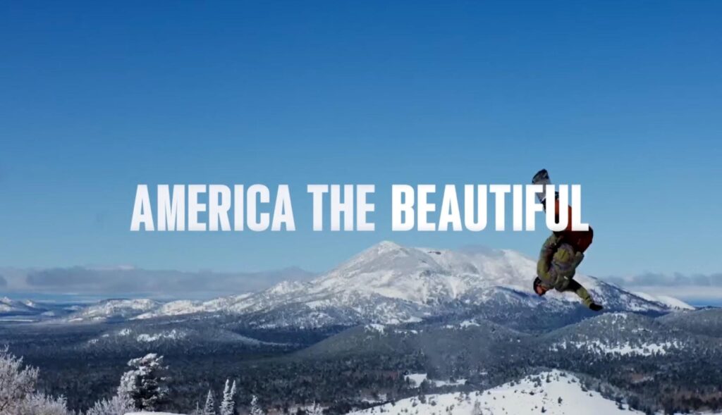 Un hombre haciendo snowboard en la nieve con las letras sobreimpresas de "America the beautiful"