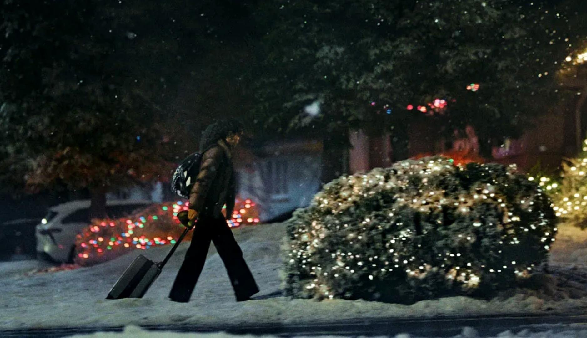 Una chica arrastrando la maleta por la Nieve en la puerta de una casa adornada con luces de Navidad