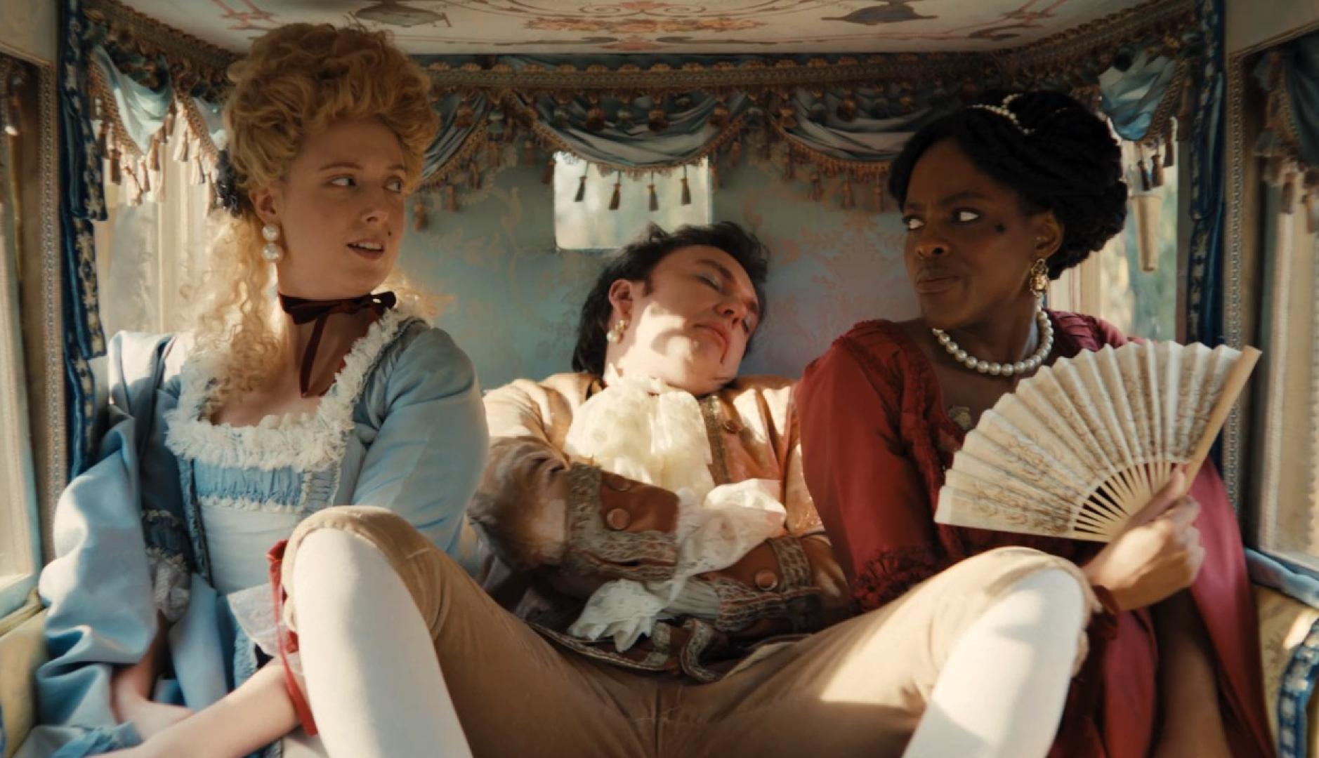 Dos mujeres vestidas de época en un carruaje con un hombre espatarrado y dormido entre ellas