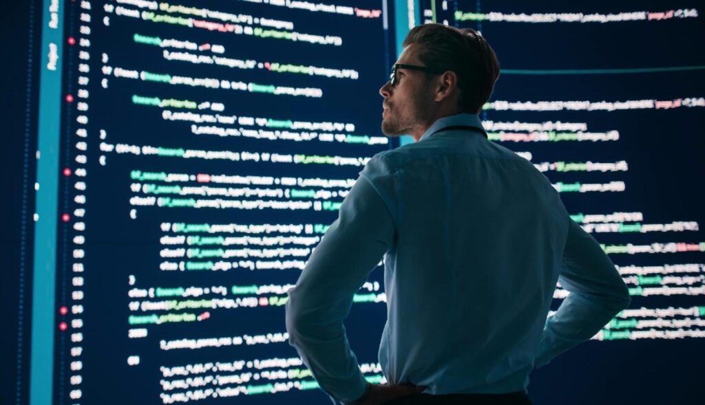 Un hombre con los brazos en jarras mira unas pantallas grandes con código HTML