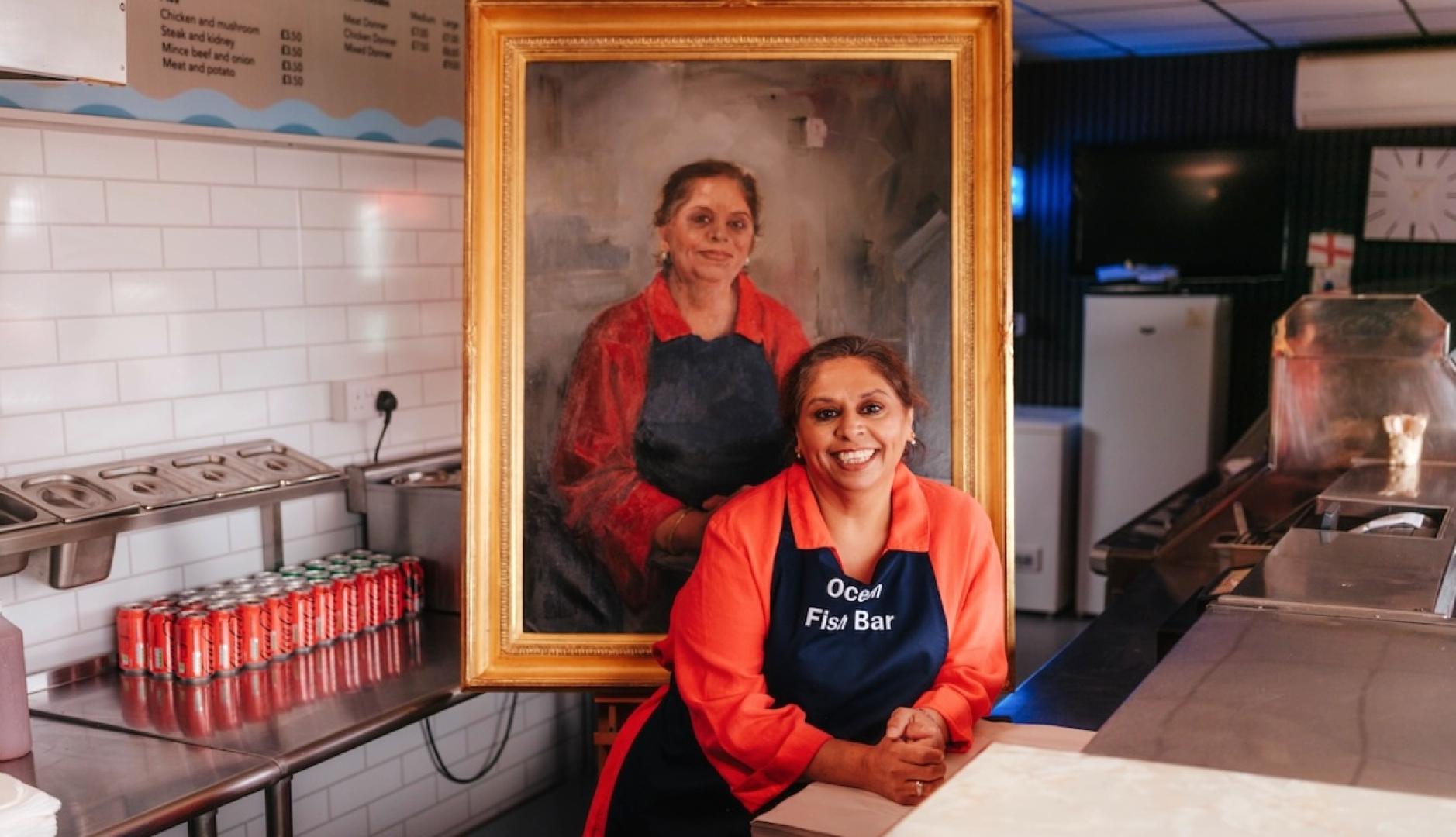 Una mujer en la cocina de su restaurante posando junto a su cuadro